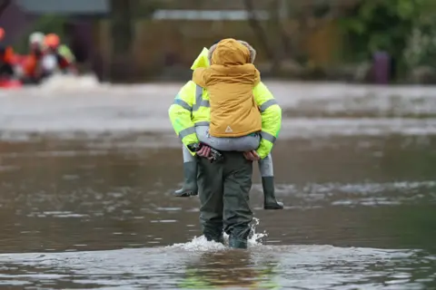 PA Media A man carries a child through floodwater in the village of Whitchurch in Herefordshire