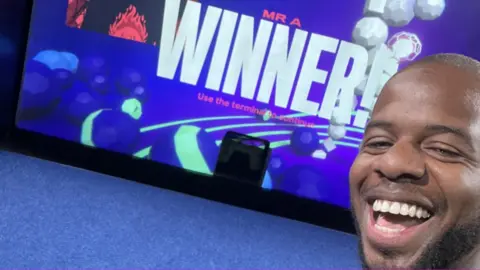 Aaron Aaron on a stage in front of a screen with the word winner