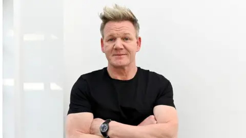 Gordon Ramsay standing against a white wall wearing a black t-shirt.