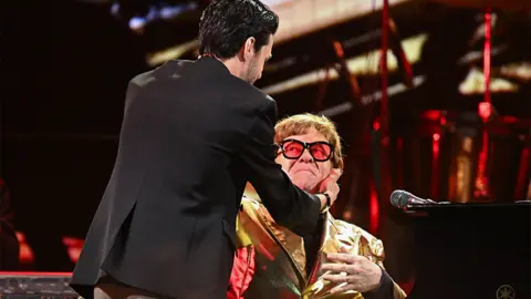 Getty Images Stephen Sanchez and Elton John embrace on stage at Glastonbury. Stephen's back is shown with Elton looking at him while sitting near his piano, with his left hand going towards Stephen. Stephen is wearing a black blazer, while Elton has red tinted glasses and a golden jacket. The background has red coloured staging