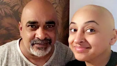 Bhandal family Sonia during her treatment with her father. They are both looking into the camera, smiling. Sonia's dad is wearing a white and grey striped top, while Sonia is wearing a black top. The background has a brown and light brown painting.