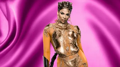 BBC/World Of Wonder Drag queen Cara Melle posing against a purple background. She has short brunette hair and is wearing big gold hoop earrings. She is wearing a skin-tight body suit with metallic armour and jewels in a bodice style at the front.
