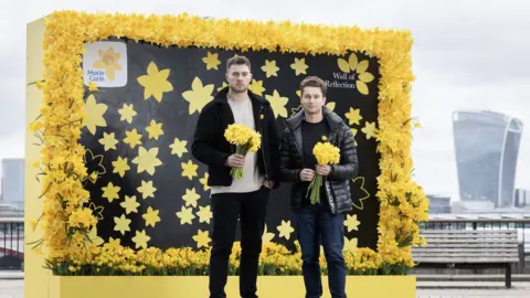 Marie Curie Curtis and AJ Pritchard hold bunches of daffodils in front of the wall on the South Bank.