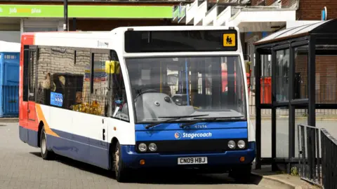 A Stagecoach bus at a bus stop. The bus is mainly white and blue but also has red and orange colours featured on the back of the bus. It is facing the camera at an angle. The bus stop is black. 