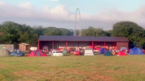 Justin Garvanovic Lots of tents can be seen in the foreground pitched in a field. Behind them is a  storage facility which has farming equipment sitting inside it. In the background there is a pylon as well as several trees.