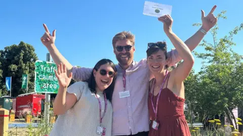 Mary-Anne O'Brien, Robin Dunlop and Laura London stood together in the sun. One of them holds a white certificate in the air and they are all smiling. Behind them are plants that form their garden show exhibition.
