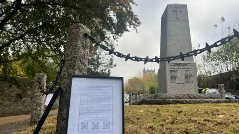 A framed picture which has names of people who lost their lives during the war leant against a concrete pillar to the left. The war memorial is on the right which has a cross at the top and names at the bottom.