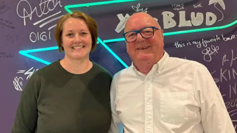 Johnny Reed/BBC A woman with short brown hair wearing a green jumper stands alongside an older bald man with glasses, wearing a white shirt. They are against a purple backdrop with handwriting across it.