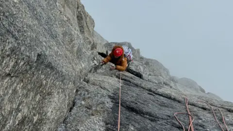 Fay Manners Tomada desde arriba, una mujer vestida de naranja, con el rostro enmascarado, sube la empinada ladera de una montaña, debajo de ella hay una espesa niebla que oscurece la caída.