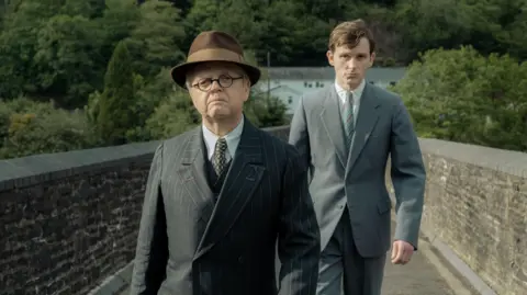 Toby Jones and Harry Lawtey in a scene in the film, walking across a footbridge with brick walls either side. They are both wearing a grey suit, white shirt and tie and Toby Jones, playing Philip Burton, wears glasses and walks ahead of Harry Lawtey 