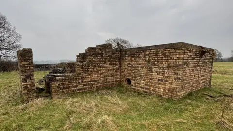 A small, brick building that has been damaged and is partially standing. There is a circular ventilation hole on one wall. It is located in a green field.