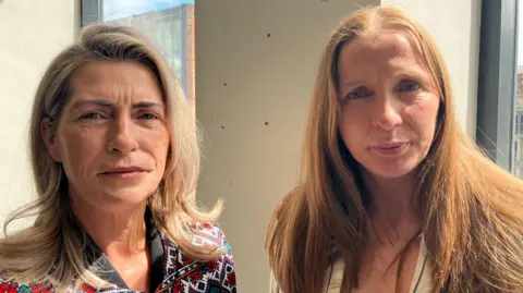 Paula, left, has shoulder length blonde hair. She is looking at the camera with a plain expression. She has a red and white patterned top on. Colleen, right, has long ginger hair. She is looking at the camera with a plain expression.