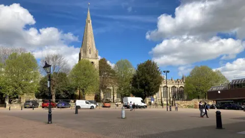 A large church is in the distance and is surrounded by trees and has cars parked in front of it. In the foreground there is a large walkway with black bollards and streetlights. There are several people walking.
