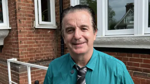 Jason Denny in a teal shirt and tie, smiling for the camera. He is stood outside a brick building with white windows.
