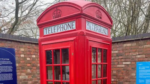 The top part of a red phone box, with the word telephone above the windows. Two rows of panes are fully visible and each row on each side of the box is divided into three sections. A wall and trees are behind the kiosk.