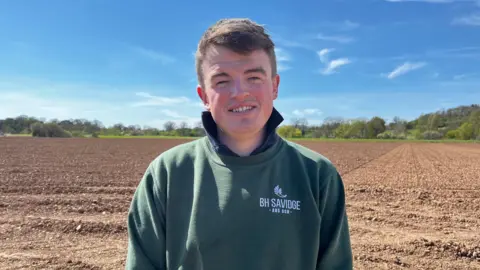 Ben Savidge stands on his farm looking at the camera. He wears a dark green jumper branded 'BH Savidge'.
