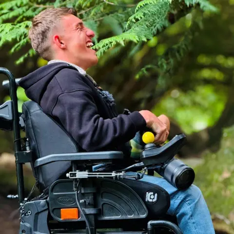 Jake Smith, a young man in a wheelchair, photographed from the side, he is laughing heartily and holding a camera.