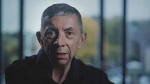 Bedfordshire Police Gary Hales is looking at the camera. He has a slim face and is clean-shaven. He has dark eyes and short, grey hair. He is wearing a dark-coloured top