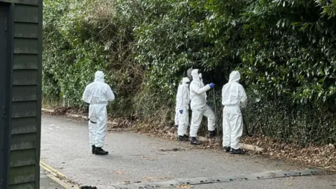 Officers in white hazmat suits search a wooded area at the side of a road. 