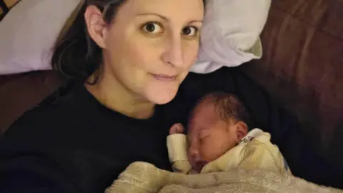 PA Media Alicia Hardie with her newborn daughter Cora. Alicia, who is wearing a black jumper and dark hair, is looking at the camera. The baby is sleeping on her mother's chest and is covered in a blanket.