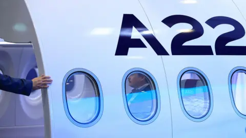 The side of an Airbus A220 plane, there are four circular windows and a blue A22 above them. There is the arm of a person in a blue jacket to the left.