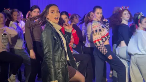 Charlotte Andrews/BBC Charlotte Steele leads a dance performance in a large room. Around 20 young female students can be seen copying her moves behind her, all wearing a variety of clothes and colours. Charlotte is standing side on and looking to the camera, wearing a large black leather jacket. She has long straight black hair and dark eyes. 