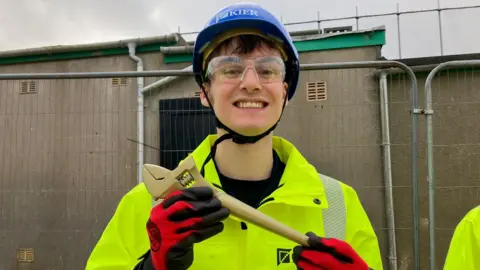 Taylor Kiltie is grinning as he holds the golden spanner. He is wearing a hard hat, safety glasses, gloves and a high-vis jacket. Behind him is metal fencing.