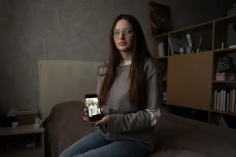 Julia Hripun sitting in her bedroom with a picture of her captive father on her iPhone.
