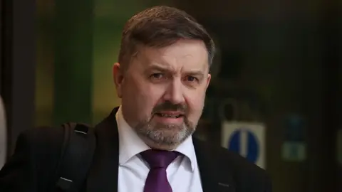 Robin Swann, who has short dark hair and beard, leaves a building wearing a dark suit jacket, a white collared shirt and a purple tie. He is carrying a black backpack strap on his right shoulder.