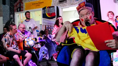 Burton Street Foundation A man in a wheelchair is wearing a pantomime style costume. It is brightly coloured in red, yellow and blue and he has exaggerated red blusher on his face. In the background are a number of people dressed in brightly coloured and sequinned outfits