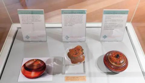St Albans Museums A display case containing three objects with information cards. On the left, there is a photograph of a round, striped container with earthy tones. In the middle, there is a fragment of an ornate, reddish-brown artefact and on the right, there is a brightly coloured, circular container with intricate red, black, and orange patterns and a small knob on top.