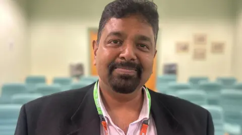 Prakash Thiagarajan in an empty lecture theatre. He has dark hair and beard, and wears a dark jacket over a pink shirt.