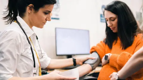A woman with dark hair tied up and a white top with lanyards around her neck is holding a blood pressure monitor on the arm of another woman with long dark hair and wearing an range top. There is a desktop computer screen behind them.