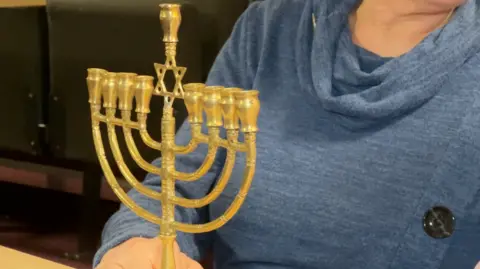 The image shows a hanukkiah on a desk being held by a woman who is sitting down. We cannot see her face. The image is zoomed in to focus on the candelabrum.