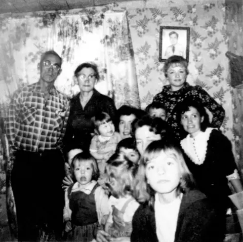 Nancy Barr/MediaPunch via Alamy Dolly Parton stands in a long sleeved patterned dress in small room with flowered wallpaper and window curtains. Above her on the wall is a slightly crooked portrait of a man. Her family, adults and children, are also crowded into the frame of the black and white image family Christmas in 1960. 