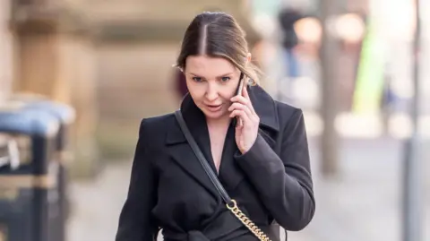 PA Media A woman wearing a black coat and a black and gold bag bows her head while holding her mobile phone to her ear