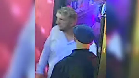 Blurry CCTV image of two middle aged men - one is wearing a black jacket and flat cap with his back to the camera, and the other is facing towards the camera, with shaggy but short greying blond hair and a blue long-sleeved shirt with the top button undone.