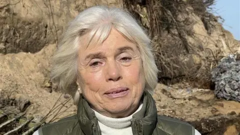 Richard Daniel/BBC Maureen Jones smiles at the camera as she stands on the coastline with a crumbling cliff behind her. She has short grey hair and wears a white turtle neck jumper with a green gilet jacket. 