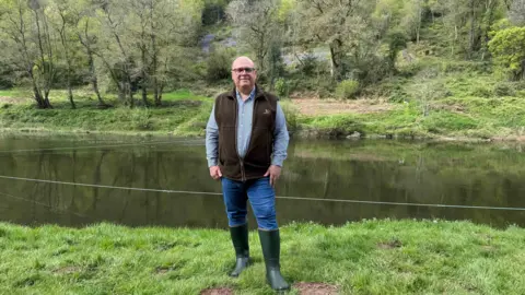 Nicola Goodwin/BBC News A fisherman stands on the banks of the River Wye