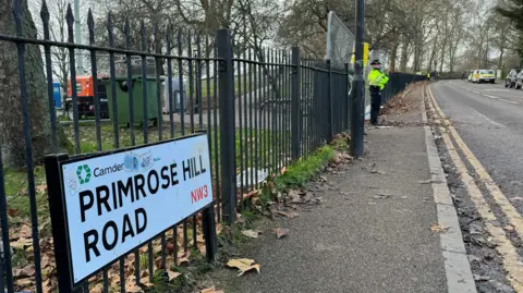 A street sign Primrose Hill Road beside railings separating the park from the pavement - in the foreground and a police officer in the background. The pavement and road 