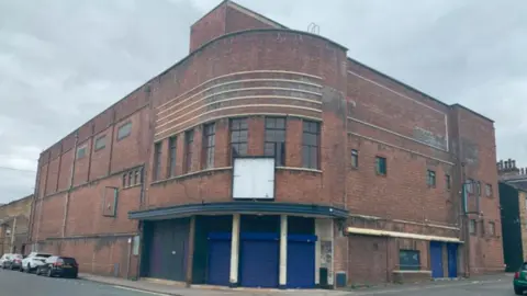 A large red‑brick corner building on a quiet street under a grey, overcast sky. The structure has a curved façade at the corner, with horizontal decorative bands and tall windows on the upper level.
On the ground floor, several blue metal roller shutters line the building, suggesting former commercial or storage use. A few cars are parked along the street to the left, and the roads around the building are empty.