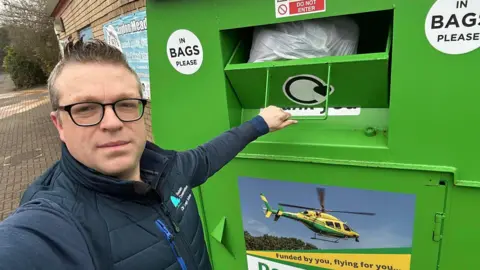 Leigh Jackson Image of a man wearing glasses, standing in front of a green clothing donation bank. In the background can be seen a brick wall and some bushes.