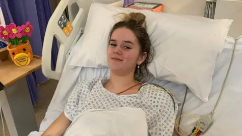 Sophia Speirs Sophia lying in a hospital bed and smiling. She is wearing a hospital gown and has a sheet over her with a number of wires coming out from underneath her gown. Her hair is tied up. A soft toy of a plant is on a table to her left. 
