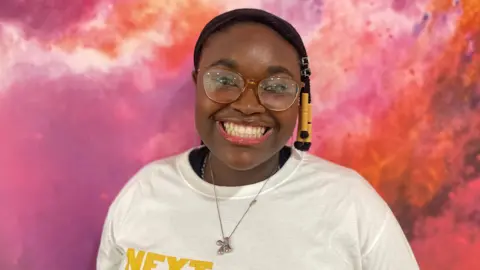 Nicola Rees/BBC Keren smiles at the camera. She has glasses and a white T-shirt saying NextGen.