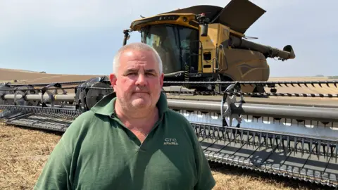 Charlie Rudler looks at the camera, wearing a green polo top. He is standing in a yellow field that has been harvested. A big combine harvester is in the background and it is a sunny day with rolling golden hills in the background.