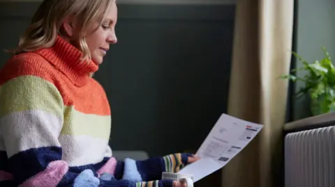 Woman sits in front of a radiator, wearing a big jumper and holding a mug and energy bill.