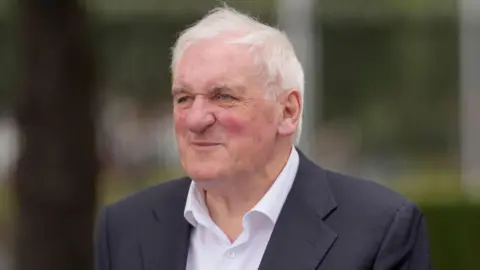 Bertie Ahern, a man with short, white hair, looks past the camera into the distance during a 2025 event. He is wearing a a navy suit jacket and a white shirt. The background is blurred