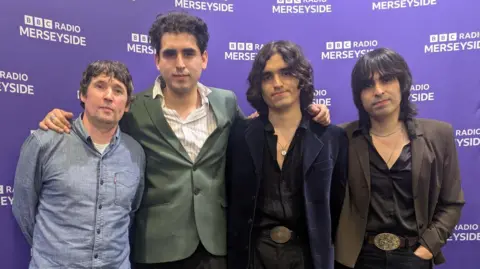 A group shot of Los Mapaches. Jevan Turner is on the left. He has short dark hair and is wearing a denim shirt. Jose Rozas has short dark curly hair and is wearing a green jacket. Fabrizio Rozas has long curly hair and is wearing an open shirt and a dark jacket. Piero Rozas has long dark hair and is wearing an open shirt and brown jacket.