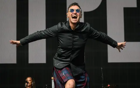 Getty Images Marti Pellow performs onstage during Radio 2 In The Park at Hylands Park in Chelmsford - he is leading forward to the crowd, arms stretched wide behind him, a roar of a smile on his face, wearing a dark red and blue kilt with lightning bolt kilt pin and a leather shirt.