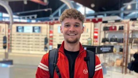 Joe is wearing a red zip up top with two white stripes on the sleeves. He is wearing it over a black top and he has a grey black backpack on. He has brown curly hair and a bit of a goatie. His eyes look brown. Behind him is bright lights with handbags and sunglasses blurred out.